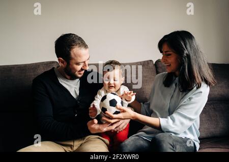 Amare i giovani genitori seduti sul divano e abbracciare carino piccolo figlio che gioca con la palla a casa Foto Stock