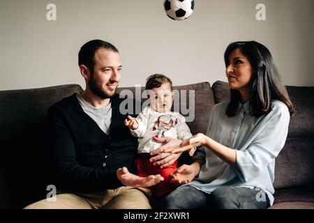 Amare i giovani genitori seduti sul divano e abbracciare carino piccolo figlio che gioca con la palla a casa Foto Stock