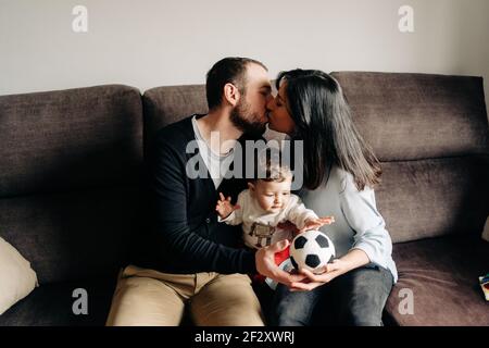 Amare il giovane uomo e la donna baciandosi mentre si siedono sul divano e abbracciando il piccolo figlio carino che gioca con la palla a casa Foto Stock