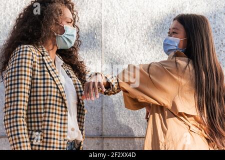 Vista laterale delle amiche etniche in abiti informali con maschere mediche che si guardano l'un l'altro mentre salutano con i gomiti Foto Stock