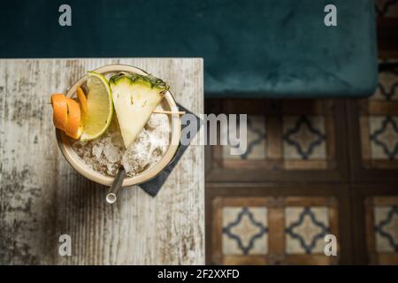 Dall'alto di una grande tiki scultorea piena di booze decorato con paglia e frutta messi su tappeto verde contro tavolo di legno Foto Stock