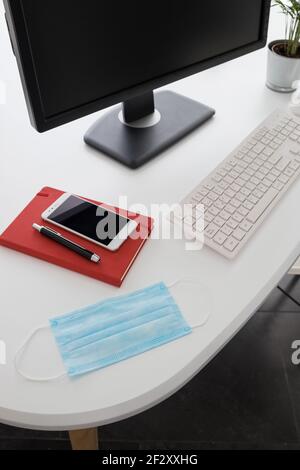 Computer e smartphone moderni con notebook posizionato su un tavolo bianco con maschera medica e pianta in vaso in ufficio leggero Foto Stock