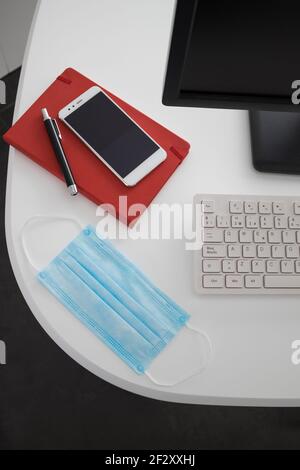 Da sopra computer e smartphone moderni con notebook posizionato sopra tavolo bianco con maschera medica in ufficio leggero Foto Stock