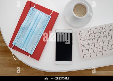 Vista dall'alto della maschera medica posizionata sul notebook e disposta con smartphone e tastiera vicino a una tazza di caffè su bianco scrivania Foto Stock