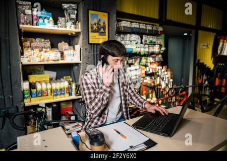 Titolare di una piccola azienda al telefono e al computer in negozio. Operaio di negozio di sport che effettua la chiamata con il telefono delle cellule. Addetto alle vendite che lavora e parla con il telefono cellulare Foto Stock