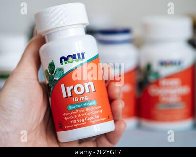 Mosca, Russia - 09 marzo 2021: Vasetto con capsule di ferro in mano femminile e altri integratori nutrizionali in sfondo sfocato. Vaso di plastica bianco e arancione ferro 18mg di Now Foods. Foto Stock