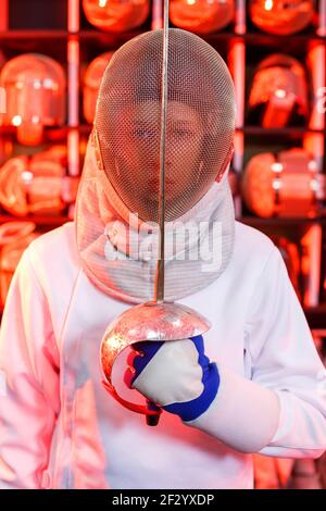 Giovane ragazzo in un vestito da scherma con una spada in mano, su uno sfondo rosso, luce al neon. L'atleta si allena. Sport, gioventù, stile di vita sano. Foto Stock