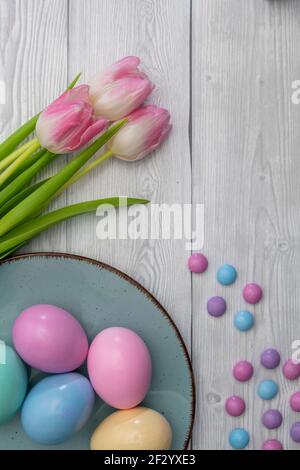 Uova di Pasqua colorate in pastello e caramelle al cioccolato colorate con tulipani bianchi rosa, su sfondo di legno bianco. Foto Stock