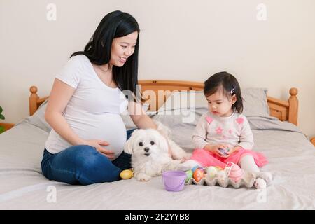 Buona Pasqua. Madre cinese asiatica incinta con la bambina che gioca con le uova di Pasqua colorate sul letto a casa. Bambino e genitore festeggiano Pasqua Foto Stock