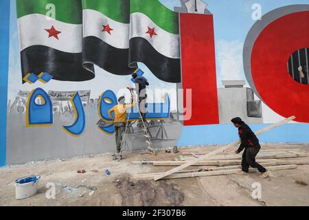 Città di Idlib, Siria. 14 Marzo 2021. L'artista Aziz al-Asmar e i suoi colleghi dipingono un murale che comprende la bandiera dell'opposizione siriana e la parola "Rivoluzione" in arabo, per commemorare 10 anni dall'inizio della guerra civile siriana, durante la quale sono state uccise almeno mezzo milione di persone, oltre un milione di feriti, mentre metà della popolazione del paese è andato in sfollamento. Credit: ANAS Alkharboutli/dpa/Alamy Live News Foto Stock