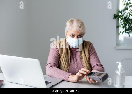 donna anziana seduta con computer portatile e terminale pos durante la quarantena Foto Stock