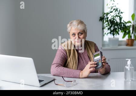 donna anziana seduta con computer portatile e terminale pos durante la quarantena Foto Stock