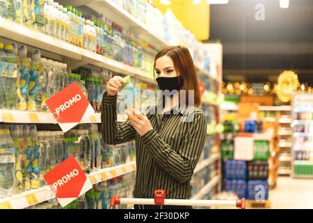 Donna con maschera nel supermercato fa acquisti di generi alimentari. Il cliente legge la composizione del prodotto nell'ipermercato. La ragazza sceglie bere. Acquirente in negozio Foto Stock