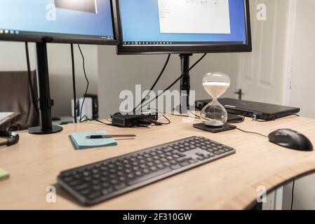 Una vista ravvicinata di una scrivania con computer, con monitor, tastiera, mouse, laptop, docking station e un timer di sabbia utilizzato come tecnica di messa a fuoco. Foto Stock