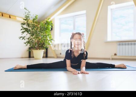 Una bambina felice in abito sportivo nero, praticando yoga, esegue una spaccatura trasversale su un tappetino ginnico, l'esercizio Samokanasana. Foto Stock