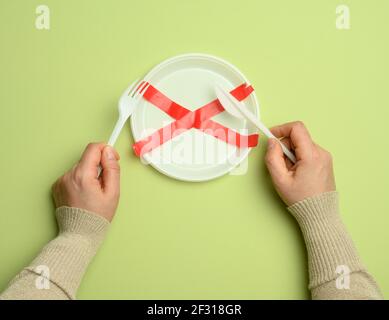 Piastra vuota in plastica bianca sigillata con nastro rosso e due mani che tengono un coltello e una forcella, sfondo verde Foto Stock