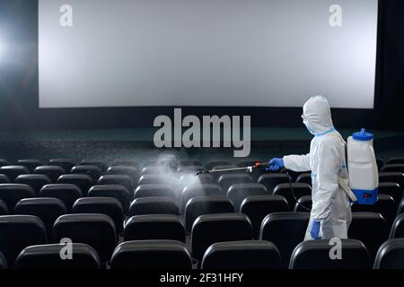 Uomo o donna in indumenti anti-virus protettivi sedili di pulizia in sala cinema con disinfettanti. Concetto di preparazione sala cinema per nuovi visitatori. Foto Stock