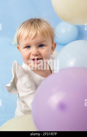 Bella ragazza allegra 1 anno con gli occhi blu in un dolcevita bianco tra palloncini e guarda il fotocamera Foto Stock