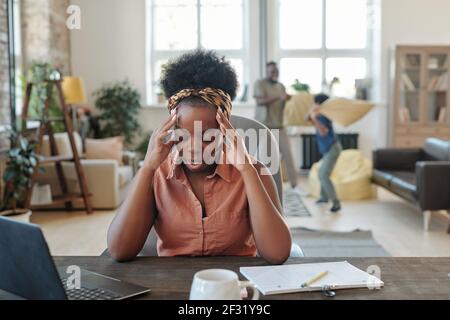 Giovane donna africana stanca che tocca la testa mentre si siede al tavolo di fronte al computer portatile durante la rete contro il marito che gioca con figlio piccolo Foto Stock