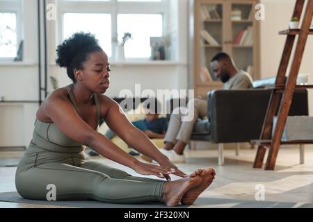 Calzare la giovane donna africana in tuta seduta sul tappetino il pavimento del soggiorno durante l'esercizio fisico contro il marito e figliolo Foto Stock