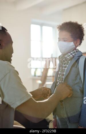 Giovane padre che abbottonava la giacca del suo figlio carino con maschera protettiva sul viso e zaino sul retro mentre vedendolo fuori a scuola Foto Stock
