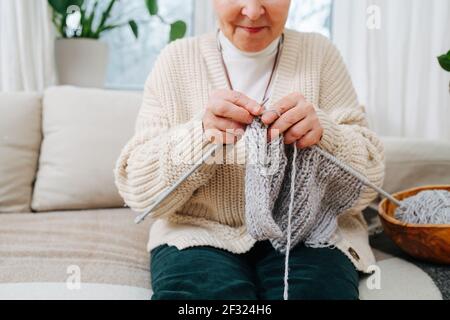 Felice granny seduto su un divano a casa, a maglia con aghi, utilizzando lana grigia. Tagliato, metà faccia. Foto Stock