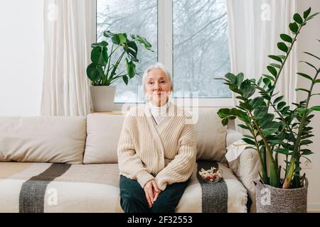 Sorridente e positivo, seduto su un divano a casa, con le mani tra le ginocchia. Dietro di lei c'è una finestra con le attrazioni invernali. Foto Stock