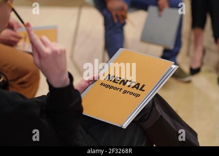 Libro o giornale del gruppo di sostegno mentale tenuto da giovani elegante donna in casualwear nero seduta di fronte allo psicologo e paziente maschile Foto Stock