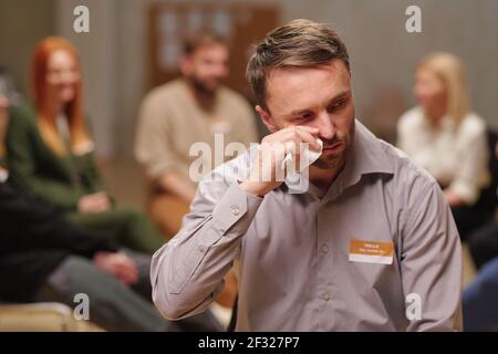 Uomo sconvolto e tormentato in casualwear strofinando le lacrime con fazzoletto contro i pazienti del gruppo di supporto mentale durante la sessione psicologica Foto Stock