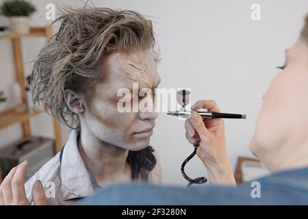 Mano di artista di visage femminile con spruzzatore elettrico di trucco che fa immagine di zombie a giovane uomo d'affari contemporaneo o attore in ufficio o studio Foto Stock