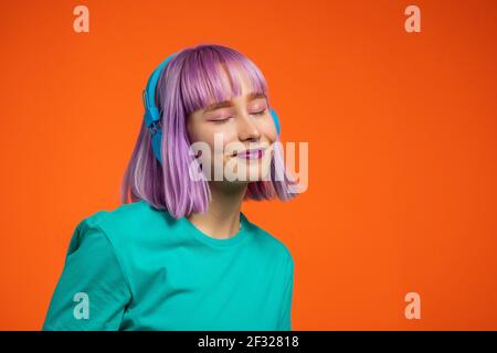 Donna eccezionale con capelli viola tinti che ascolta musica in cuffia e canta su sfondo arancione. Unica ragazza hipster danzare Foto Stock
