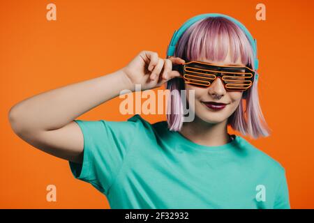 Donna eccezionale con capelli viola tinti che ascolta musica in cuffia e canta su sfondo arancione. Unica ragazza hipster danzare con reticolo Foto Stock