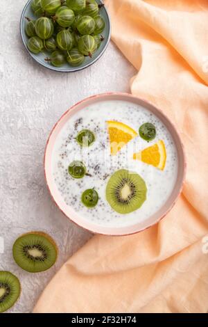 Yogurt con kiwi, uva spina, ciotola in ceramica su fondo grigio in cemento e tessuto di lino arancione. Vista dall'alto, piatto, primo piano. Foto Stock