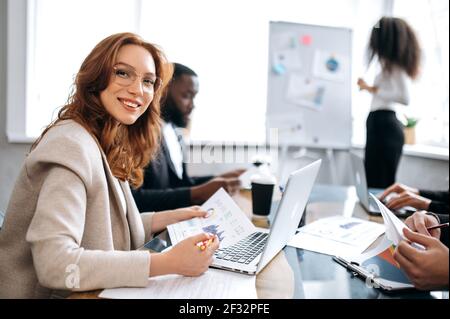 Una giovane e attraente donna d'affari è seduta alla scrivania della conferenza, utilizzando un computer portatile. Bella donna dipendente in riunione briefing, facendo documenti, guardando la fotocamera, sorriso Foto Stock