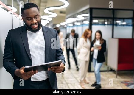 Uomo d'affari influente in abito elegante formale sta guardando il tablet, sorridendo, navigare in Internet. Un impiegato africano di successo è in piedi nella sala d'ufficio, lavorando al progetto Foto Stock