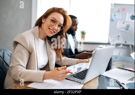 Bella donna rossa sta usando il laptop, lavorando al progetto con i colleghi. Una donna felice in tuta elegante è seduta in un ufficio moderno durante una riunione d'affari, guardando la fotocamera, sorridendo Foto Stock