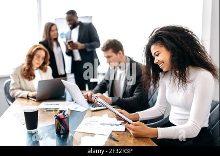 Felice attraente dipendente femminile è seduto in ufficio a riunione di affari, imparando grafici finanziari. Bella giovane donna di affari adulta facendo documenti, sorridente Foto Stock