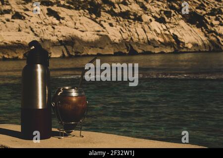 Mate y termo en la playa- bere Mate sulle rocce. Spiaggia mediterranea con acque turqoise trasparenti. Buona sensazione. Stile di vita Helthy Foto Stock