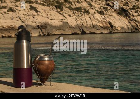 Mate y termo en la playa- bere Mate sulle rocce. Spiaggia mediterranea con acque turqoise trasparenti. Buona sensazione. Stile di vita Helthy Foto Stock