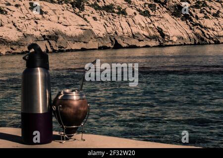 Mate y termo en la playa- bere Mate sulle rocce. Spiaggia mediterranea con acque turqoise trasparenti. Buona sensazione. Stile di vita Helthy Foto Stock