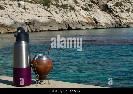 Mate y termo en la playa- bere Mate sulle rocce. Spiaggia mediterranea con acque turqoise trasparenti. Buona sensazione. Stile di vita Helthy Foto Stock