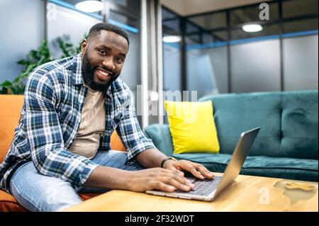 Allegro ed elegante freelancer seduto sul divano, utilizzando un computer portatile. Buon programmatore afro-americano guardando la telecamera, sorridendo, lavorando su un nuovo progetto o app Foto Stock