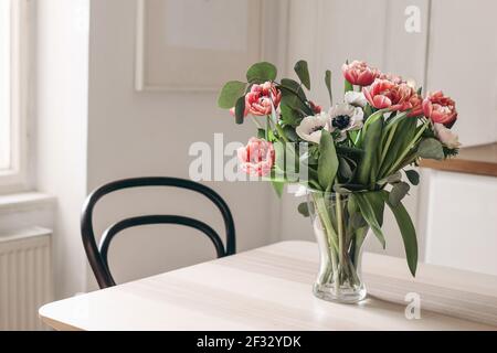 Fiori primaverili in vaso di vetro su tavolo di legno. Sfondo della cucina sfocato con sedia vecchia. Bouquet di tulipani rossi, fiori bianchi di anemone ed eucalipto Foto Stock