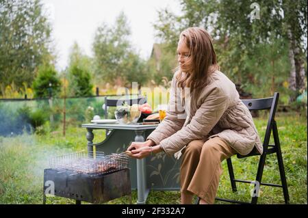 Una giovane ragazza ottimista sta preparando un barbecue per tutta la famiglia nel cortile del palazzo di famiglia. Vacanze autunnali in auto-isolamento. Foto Stock