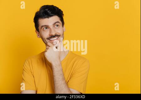 Ritratto di un bel ragazzo caucasico ottimista vestito in t-shirt arancione, guardando penosamente verso lo spazio vuoto, pensando nuove idee, sorridendo, si erge su isolato sfondo arancione Foto Stock