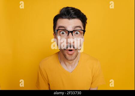 Ritratto di stupido allegro ragazzo caucasico in occhiali e in una t-shirt arancione, allegro guardando la fotocamera con un volto distorto, in piedi su uno sfondo arancione isolato Foto Stock