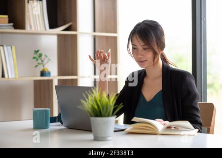 Donna d'affari asiatica che parla con il team di colleghi in videochiamata conferenza nota di scrittura sul libro con la faccia del sorriso Foto Stock