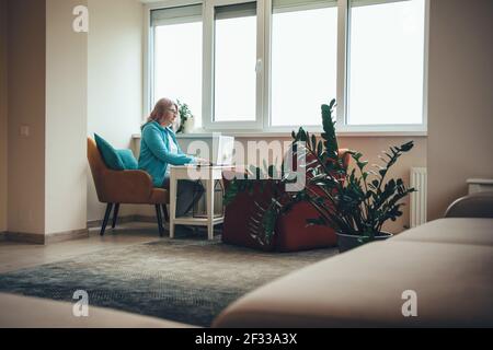 Donna caucasica senior è concentrata sul lavoro da casa utilizzando un computer portatile vicino alla finestra Foto Stock