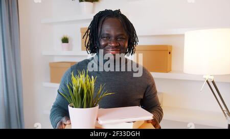 Eccitato giovane afroamericano che si sposta nel nuovo appartamento. Scatole di cartone con piante e libri. Foto di alta qualità Foto Stock