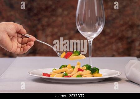 Fuoco selettivo di una forchetta con pasta e verdure tenuto una mano su un piatto di insalata di pasta su uno sfondo fuori fuoco. Concetto di cibo sano. Foto Stock